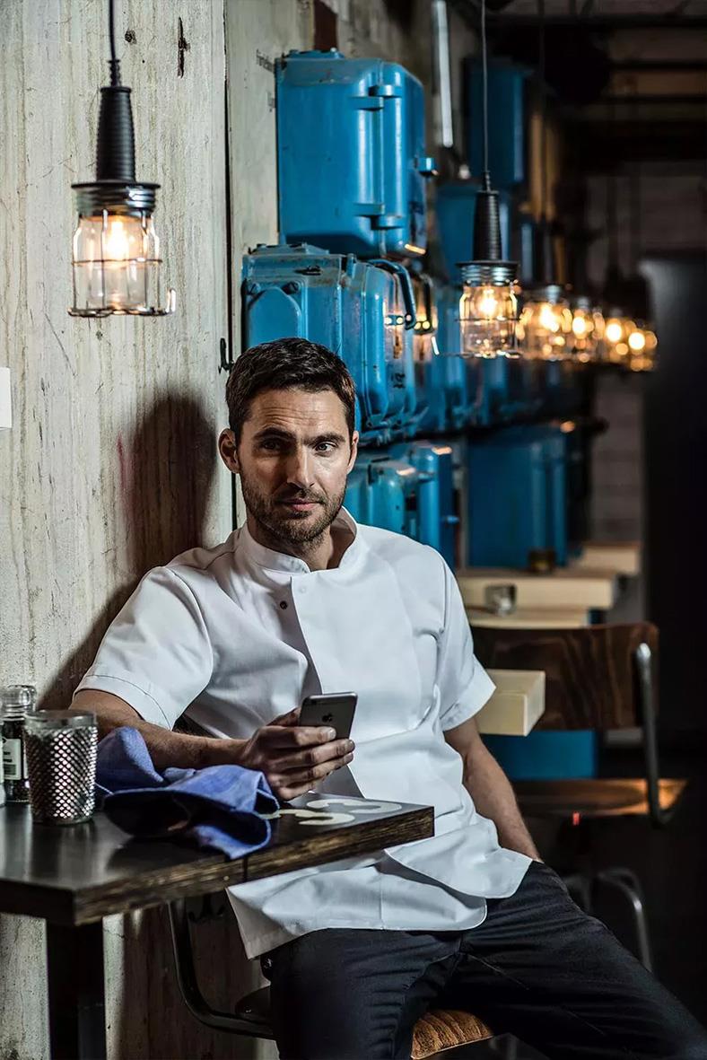 Kock i vit uniform sitter i en restaurang med trävägg och blå maskiner i bakgrunden, tittar på sin telefon
