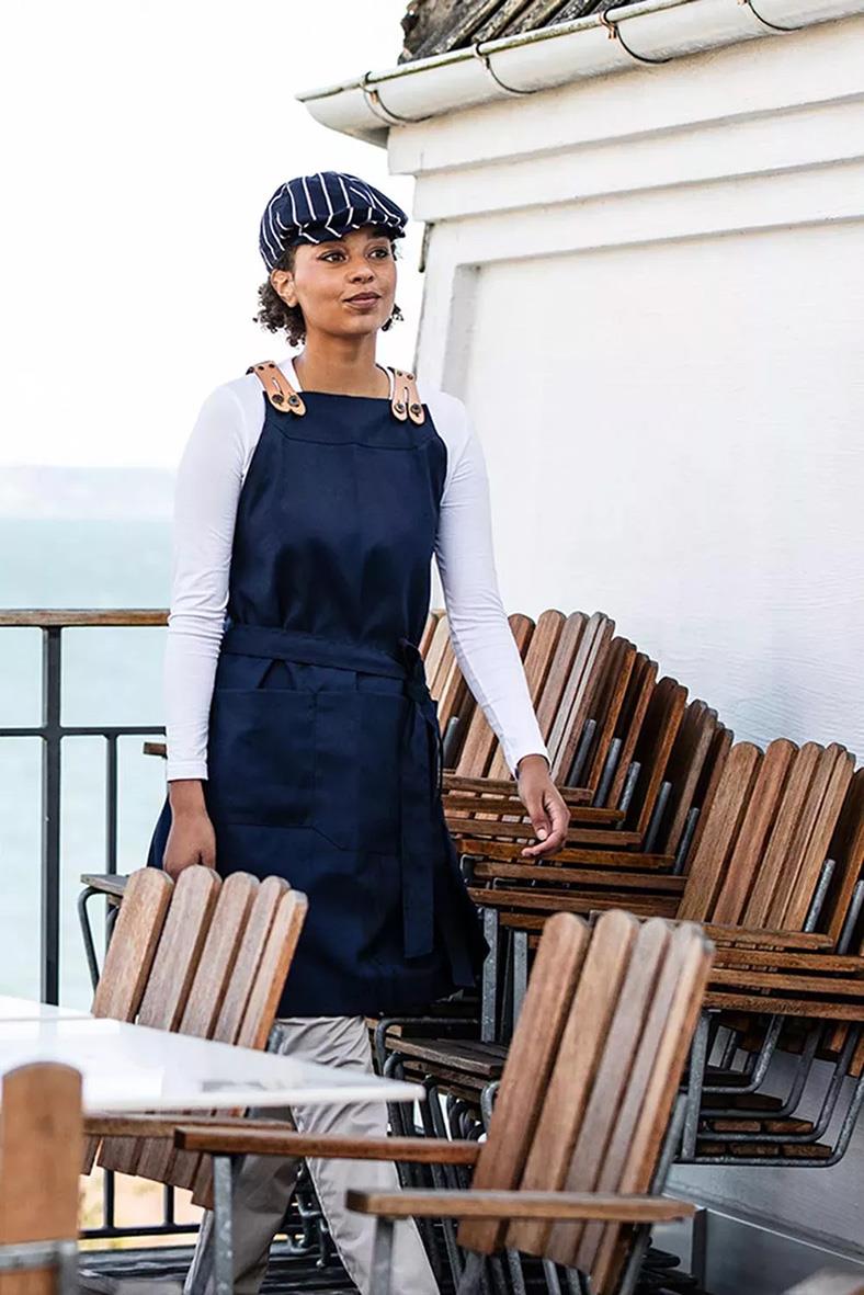 Kvinna i marin förkläde och strumpor, stående på uteservering med trästolar och utsikt över havet