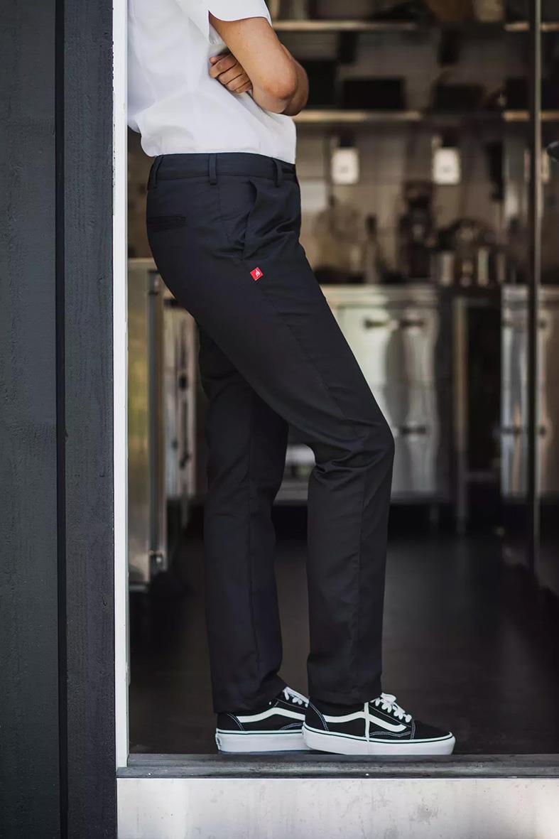 Person i svarta midjebyxor och vit t-shirt står vid en vägg i ett kök, med armar korsade och sneakers på golvet.