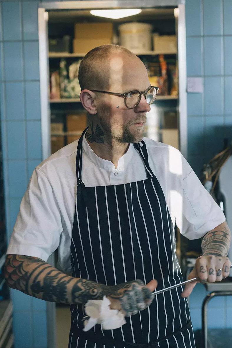 Kockpiké Segers 6109 Unisex vit 3XL, man med tatueringar, glasögon, vit kockskjorta och svartvit randig förkläde, står i kök