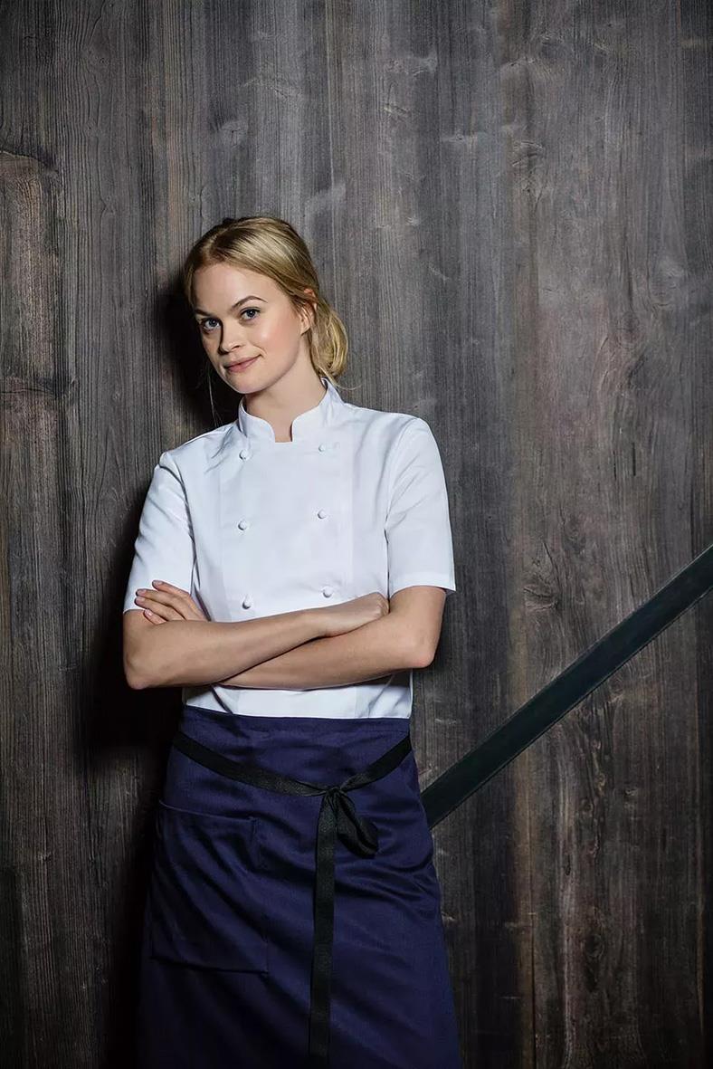 Kockklädd kvinna i vit kockrock och mörkblå förkläde står mot en trävägg, korsar armarna och ler lätt.