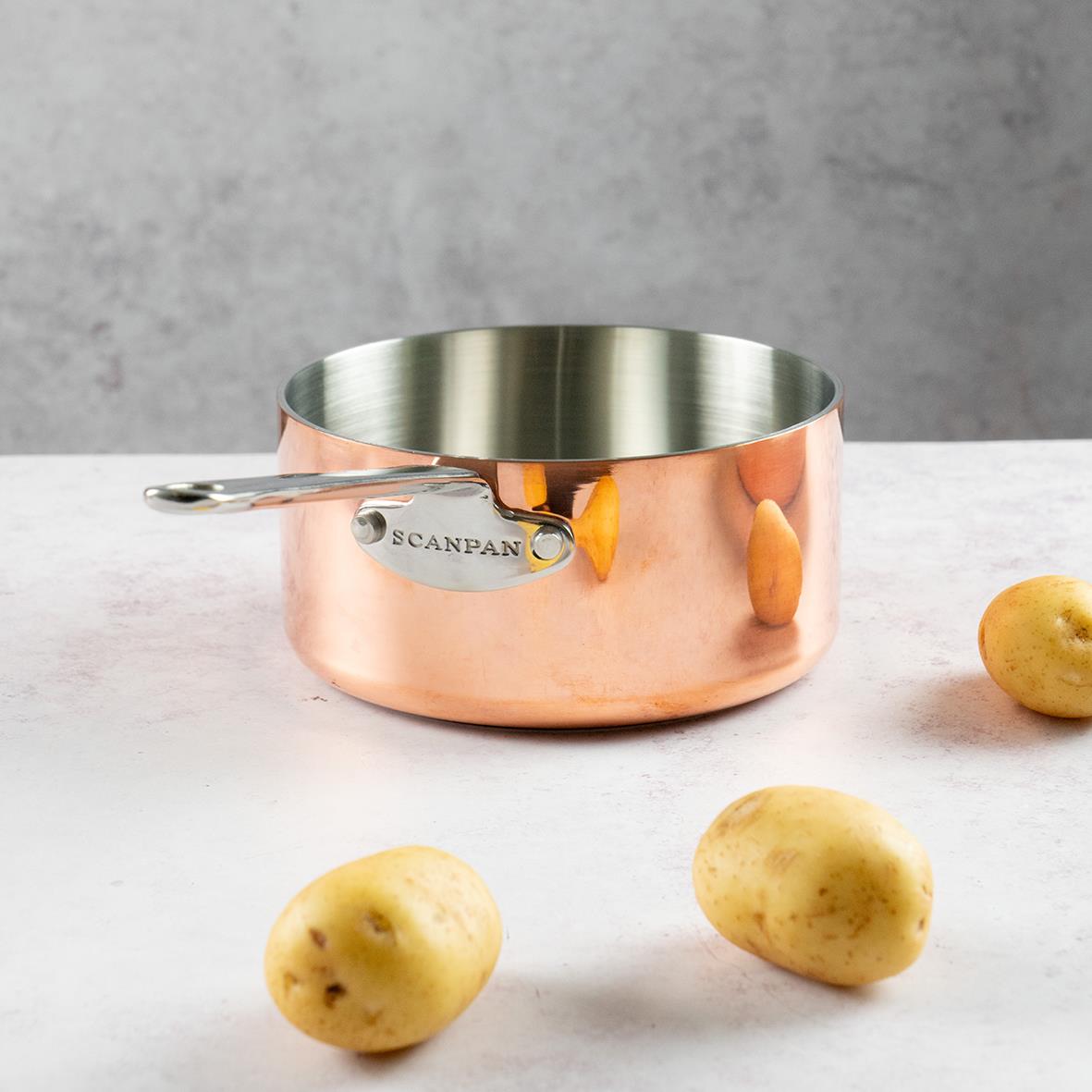 Kastrull Scanpan Maitre D’ Koppar Ø16cm 1,5L med tre potatisar framför på en ljus yta
