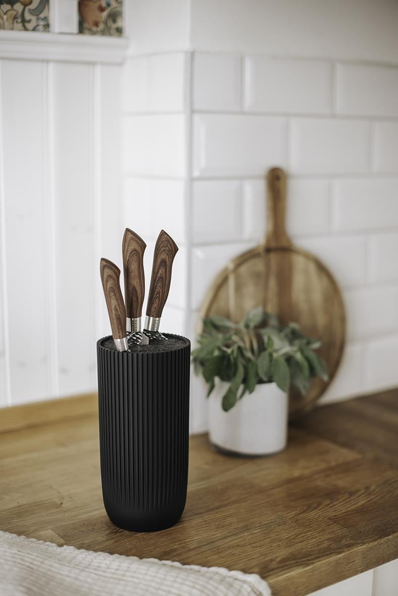 Svart knivställ med tre knivar med trähandtag på ett köksbord, bakgrund av vit vägg och köksredskap