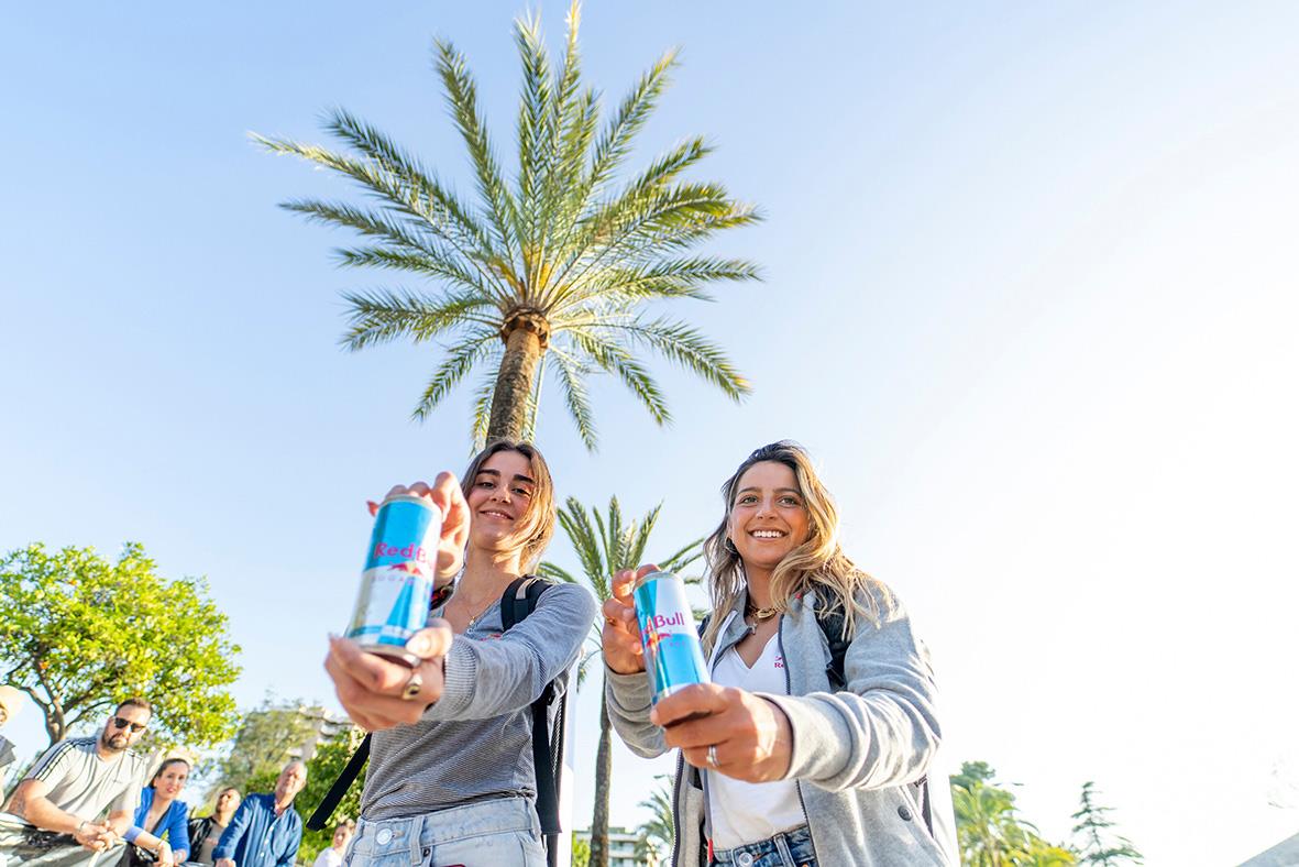 Två unga kvinnor som håller Red Bull sockerfri energidryck i en park med palmträd och andra människor i bakgrunden