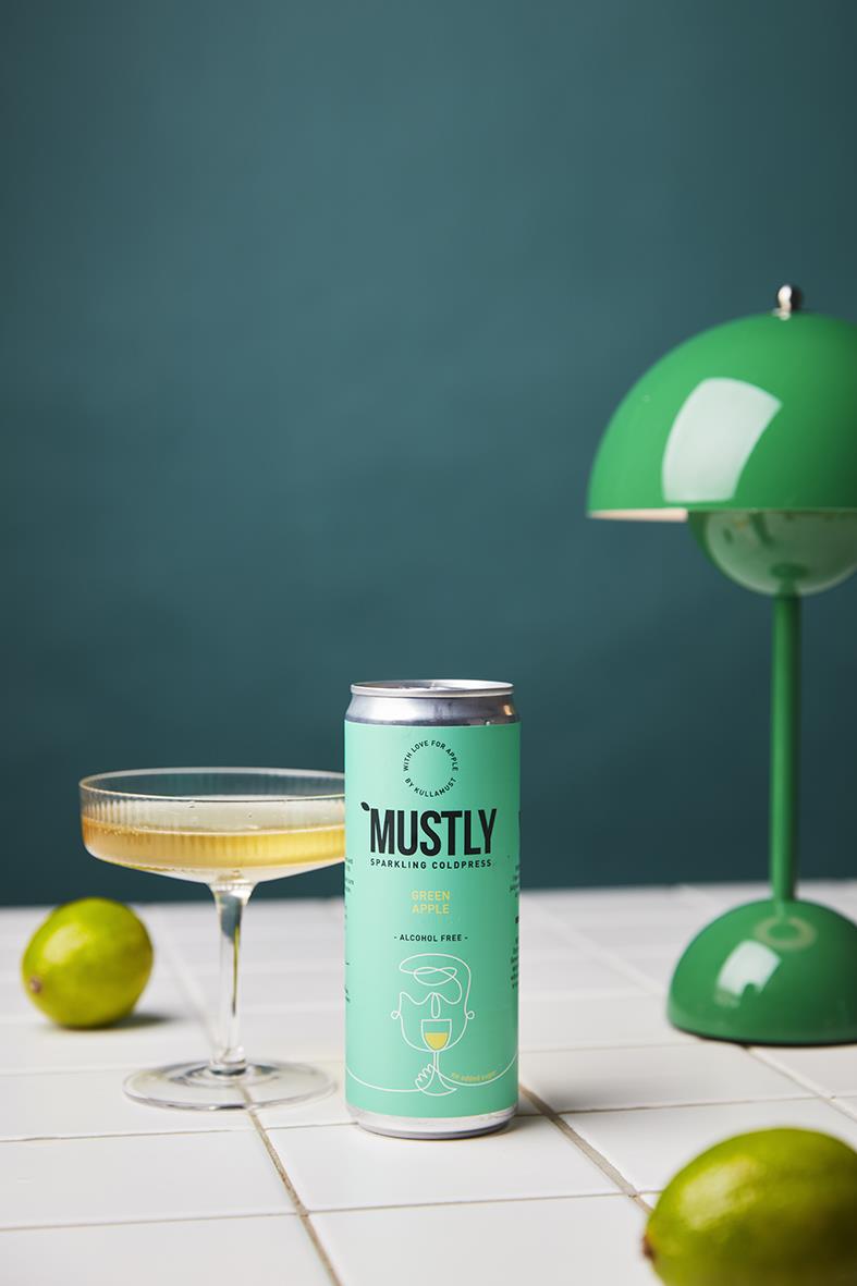 Grön burk av Mustly grönt äpple dryck 33cl med två gröna äpplen och en grön bordslampa på en vit bänk
