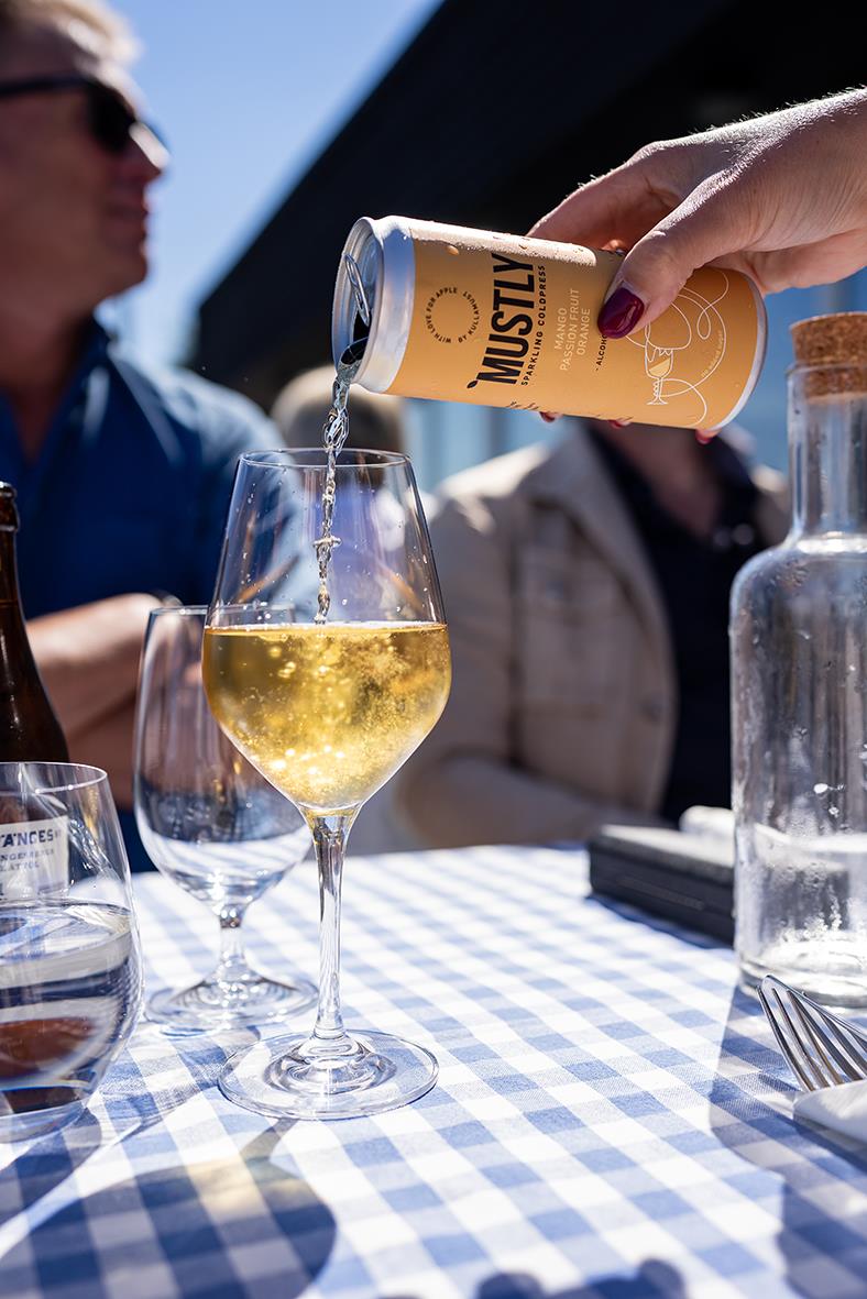 Person häller Mustly Mango&Passion dryck i ett glas på ett utomhusbord med andra glas och en vattenkanna