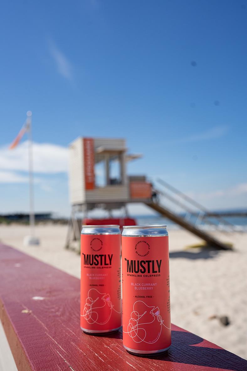 Två burkar Mustly blåbärsdryck på en strandbänk med badtorn och en strandhytte i bakgrunden