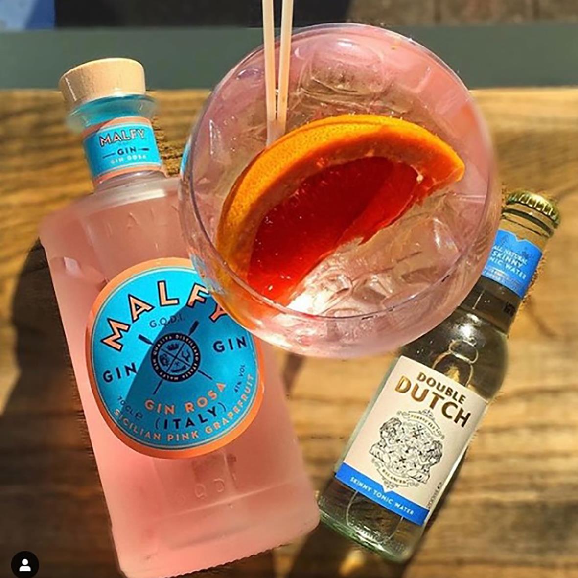 En glas med rosa drink, apelsinskivor och sugrör, omgiven av ginflaskor på ett träbord