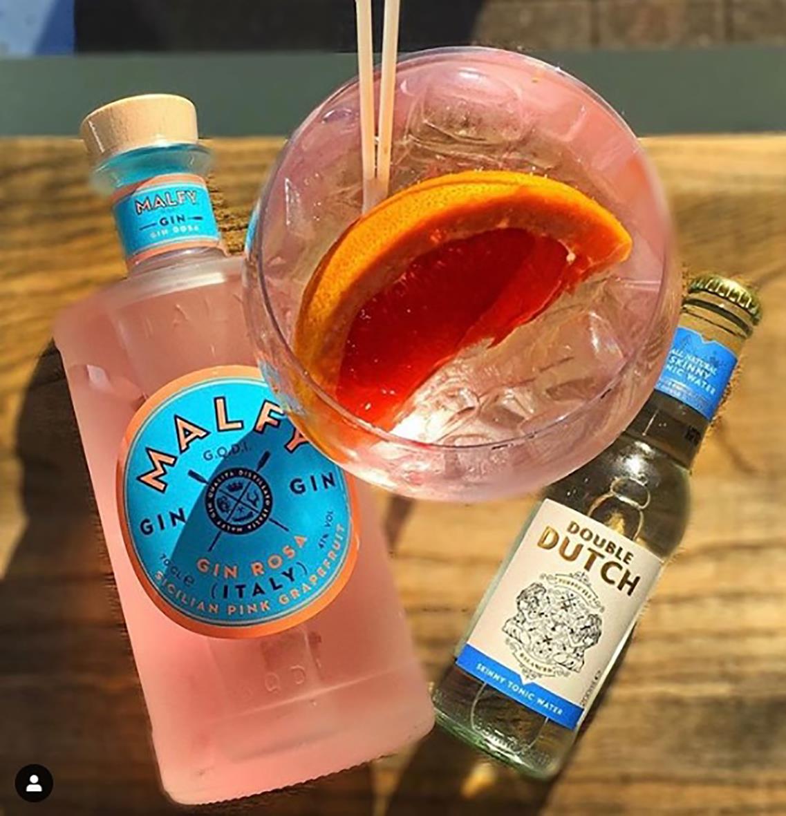 En glas med rosa drink, apelsinskivor och sugrör, omgiven av ginflaskor på ett träbord