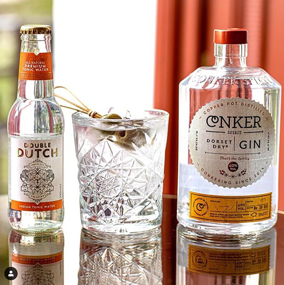 Dubbel Dutch Tonic Indian 20cl, en glas med is och en liten sked, samt en ginflaska på ett bord med röd bakgrund