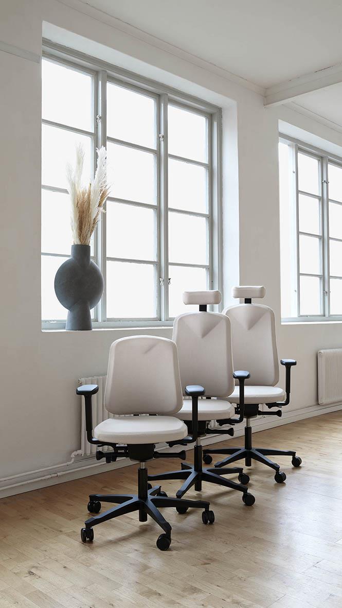 Tre vita kontorsstolar med nackstöd och svarta hjul framför stora fönster i ett ljust rum
