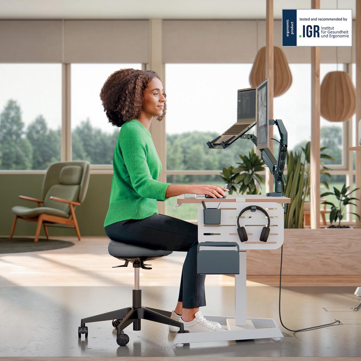 Kvinna sitter på svart ergonomisk kontorsstol med hjul framför skrivbord med dator och hörlurar i ett modernt kontorsutrymme