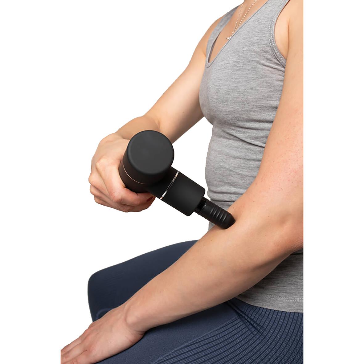 Person använder massagepistol på överarm, grått träningslinne och blåa träningsbyxor