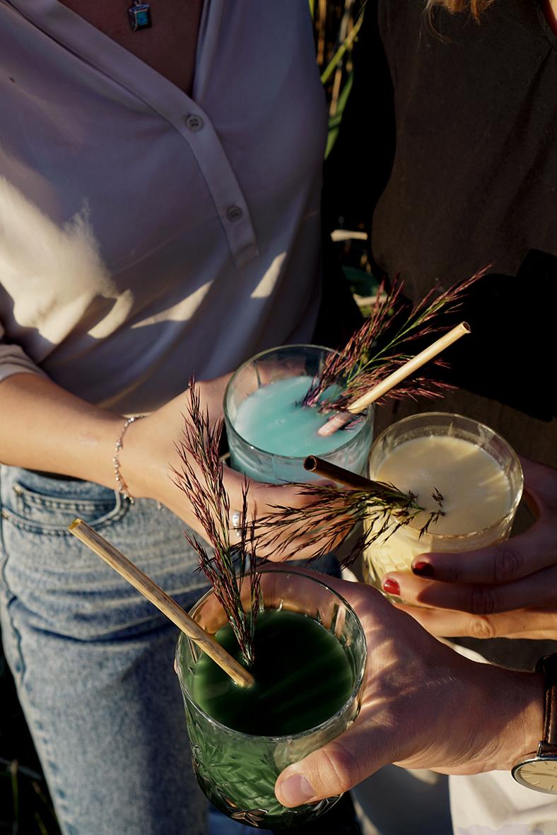 Tre personer håller i tre färgglada drinkar med sugrör och dekorationer, utomhus i solsken.