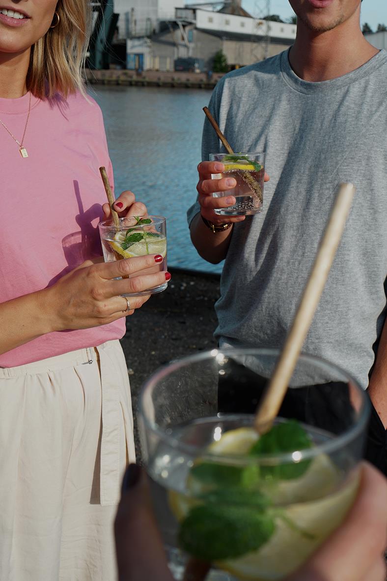 Två personer håller drinkar med sugrör, bakgrund av vatten och båtar, i sommarmiljö