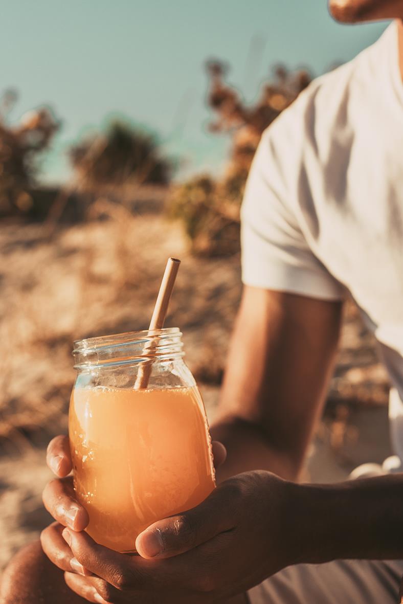 Person håller ett glas med juice och ett sugrör utomhus, bakgrund av natur och klar himmel
