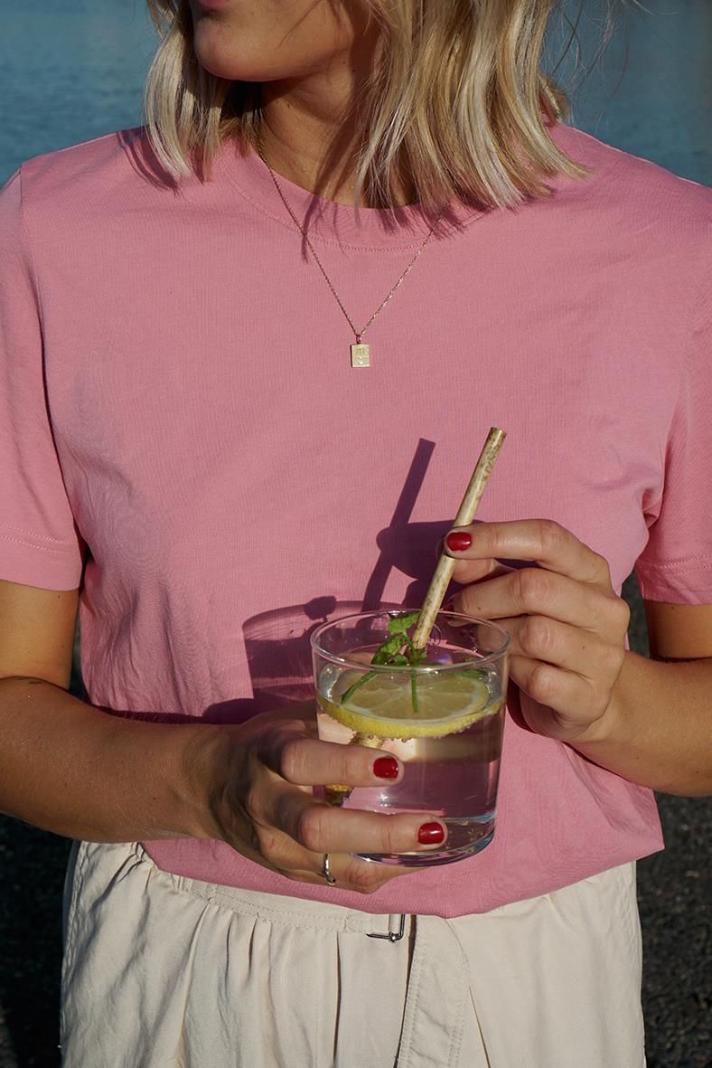 Kvinna i rosa t-shirt håller ett glas med vatten och ett sugrör av trä, utomhus