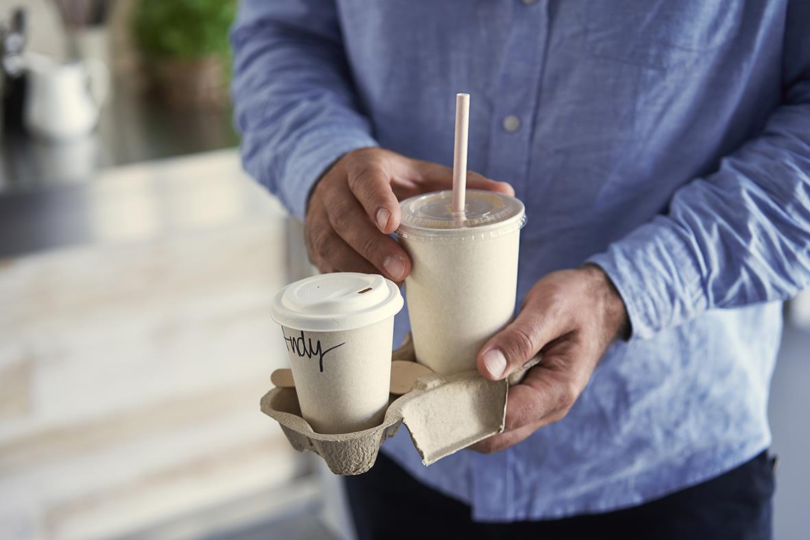 Person håller en pappersmugg och en större kopp med sugrör, båda i Duni Breeze Bagasse/PLA naturmaterial
