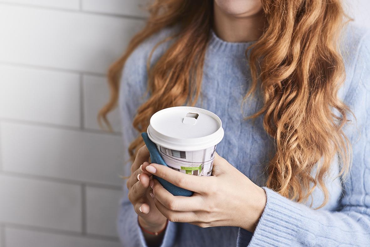 Kvinna håller en kopp med lock och papper i handen, klädd i blå tröja, mot vit vägg