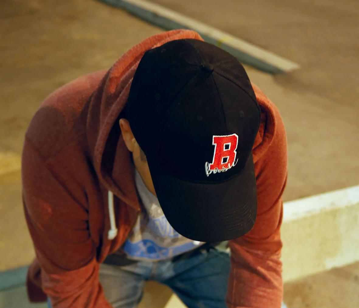 Person bär svart keps med röd B-logga, orange hoodie och blå jeans, sittande på marken