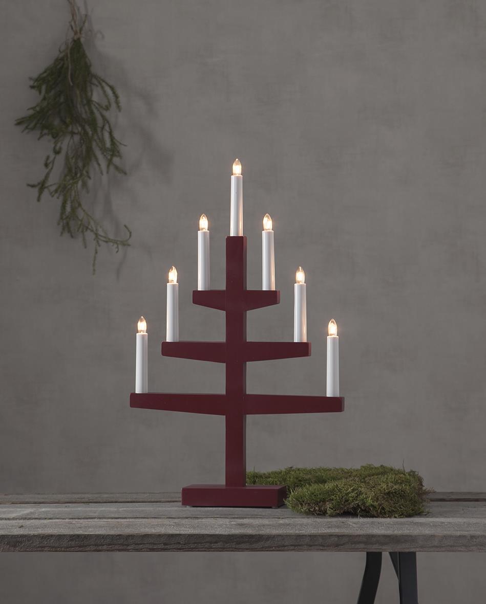 Röd ljusstake i form av ett julträd med sju ljus, placerad på ett bord med mossa och en grön kvist i bakgrunden