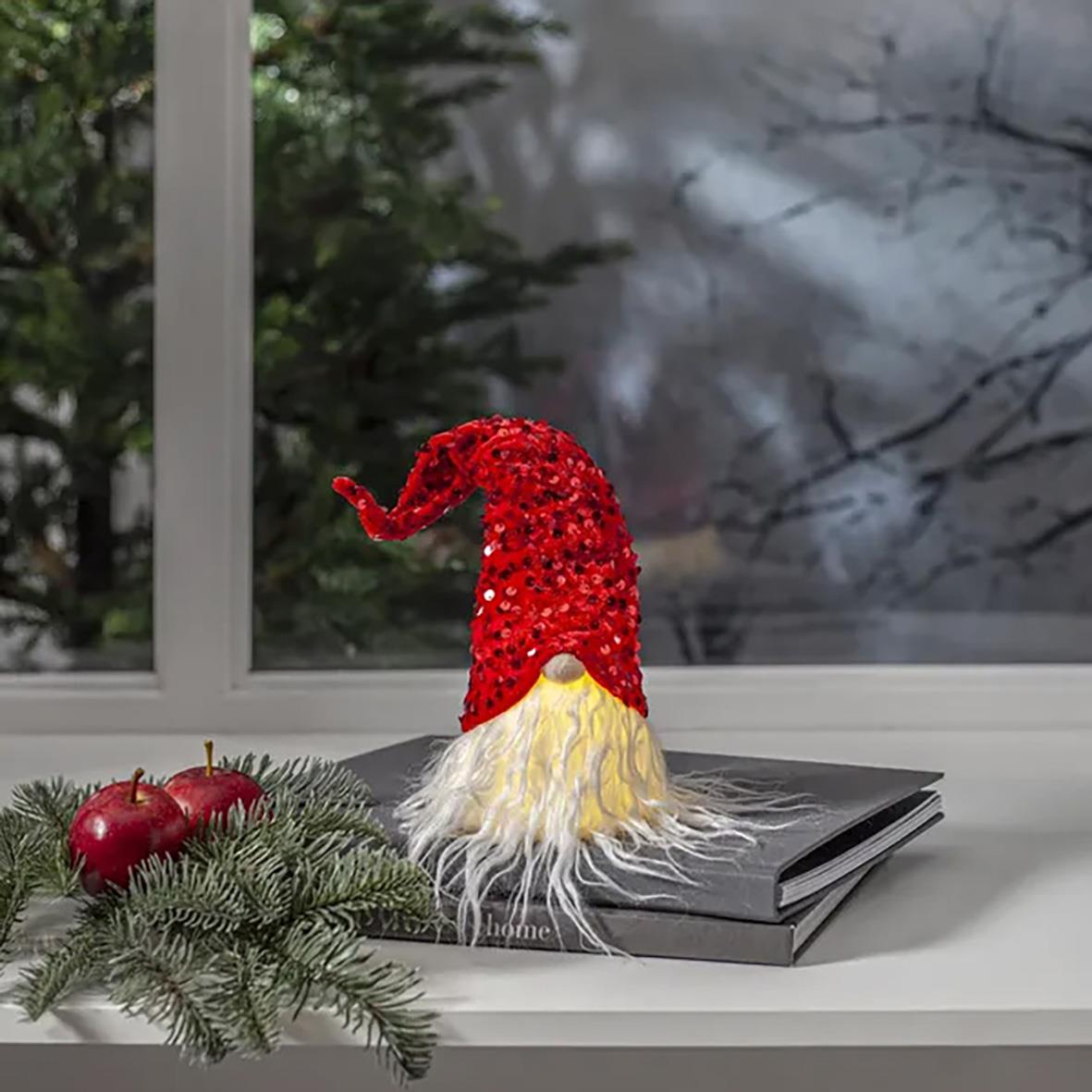 Dekorationsfigur tomte med röd luva och långt vitt skägg på fönsterbräda med julgransgrenar och röda äpplen