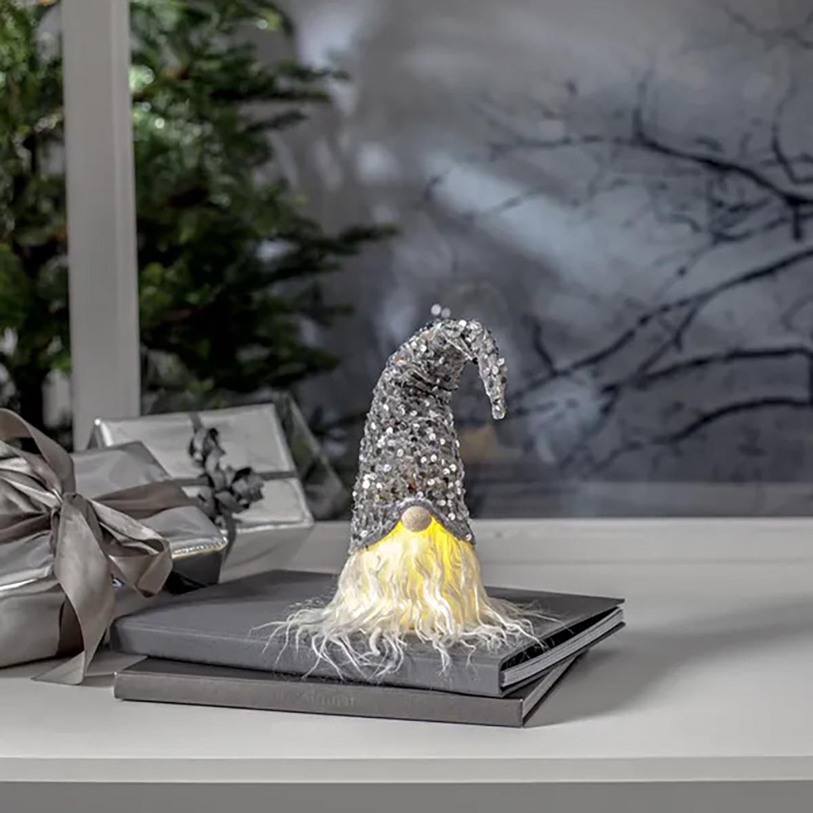 Snygg tomtefigur med silverglitterhatt och vit skägg på en vit yta med julgranar och presenter i bakgrunden