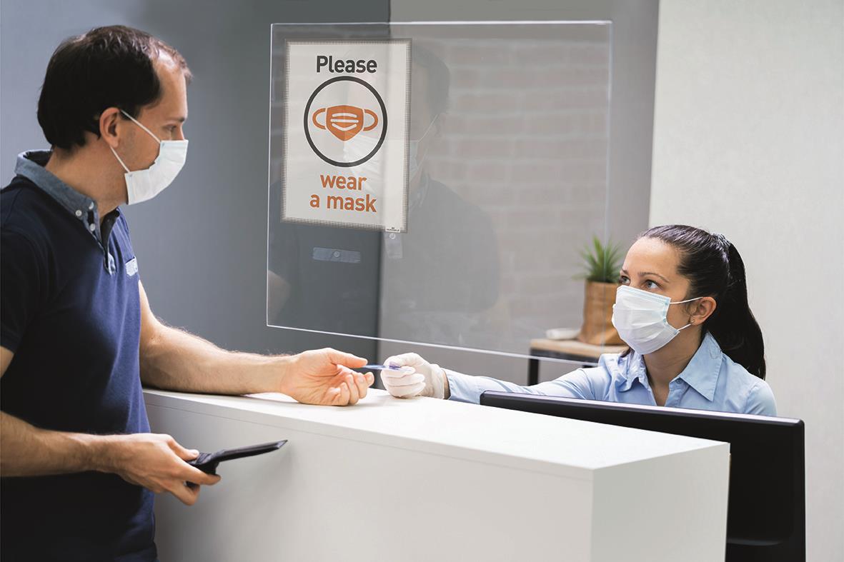 Person som köper en grå A4 infoficka av plast från en receptionist bakom en plexiglasvägg, båda bär ansiktsmasker.