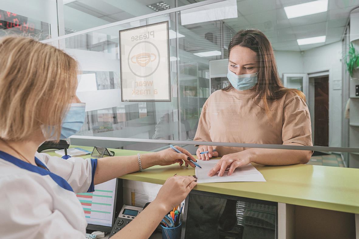 Kvinna i ansiktsmask som lämnar in en blankett till en annan kvinna bakom en glasruta i ett kontorsmiljö