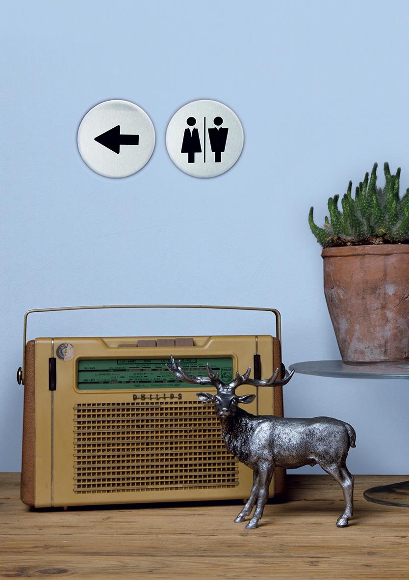 Dörrskylt med pilar och symboler för kvinnor och män, rostfritt stål, 10x10cm, placerad på vägg ovanför en gammal radio och en växt