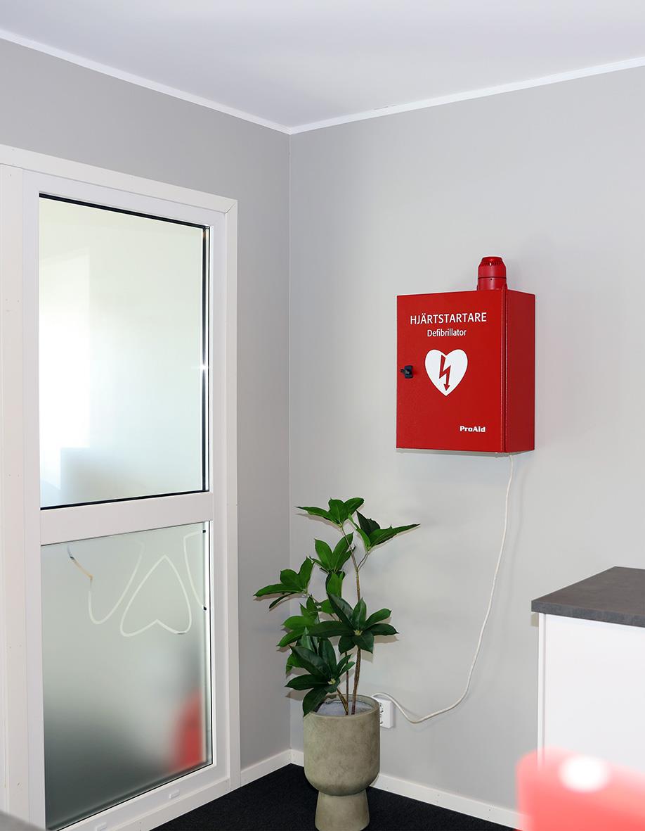 Röd skåp för hjärtstartare med SMS-larm, placerad på vit vägg bredvid fönster, med växt framför.