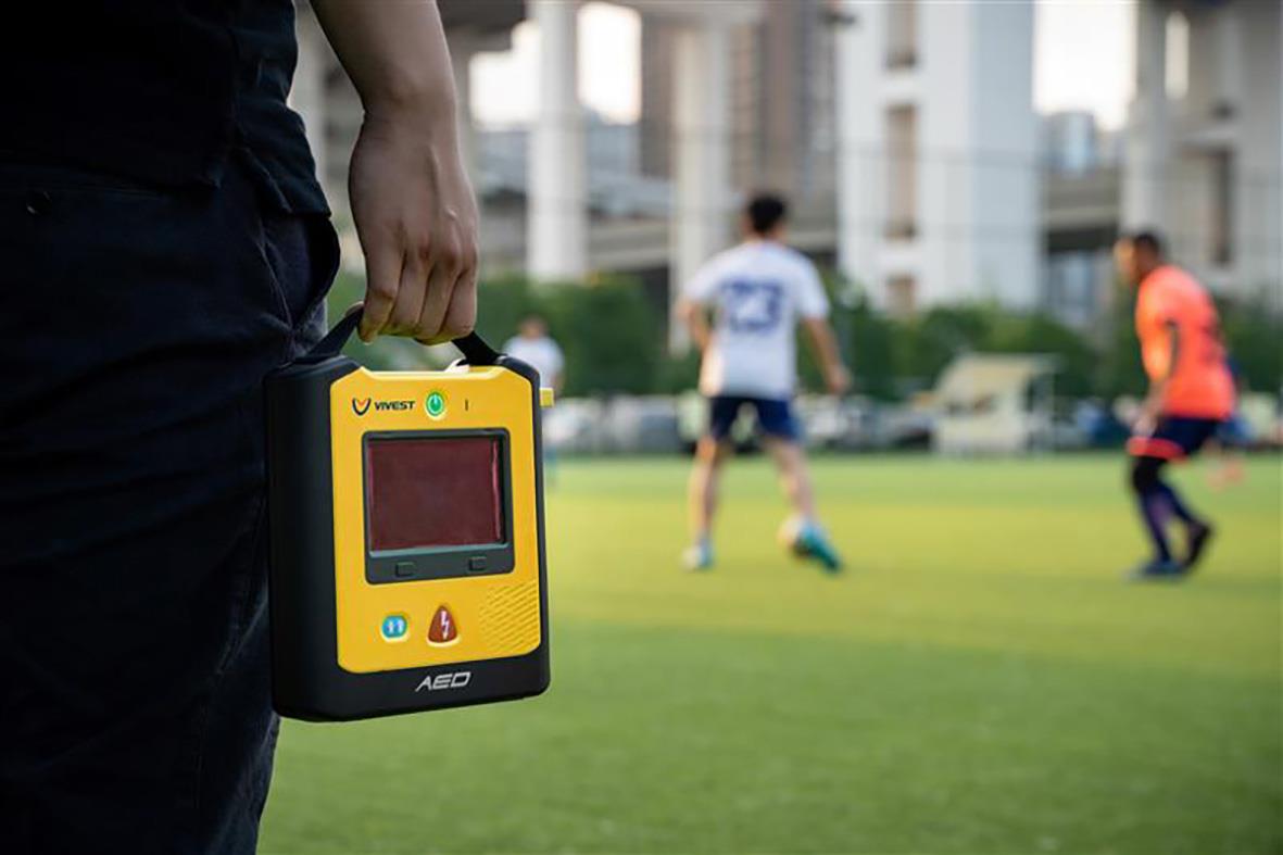 Person håller en hjärtstartare på en fotbollsplan med barn som spelar i bakgrunden