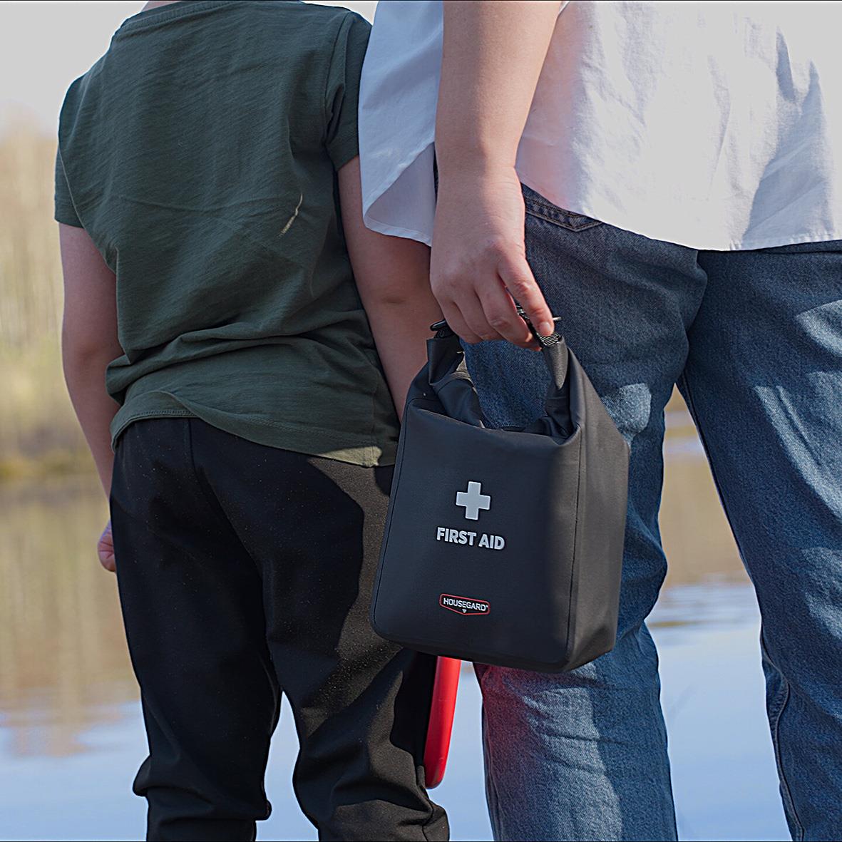 Två personer står vid vatten, en håller en vattentät Första Hjälpen-väska från Housegard
