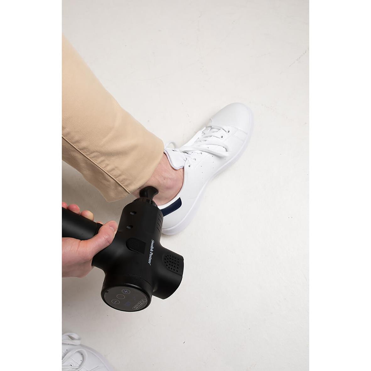 Person använder massagepistol på foten i ett rum med vit golv och beige byxor