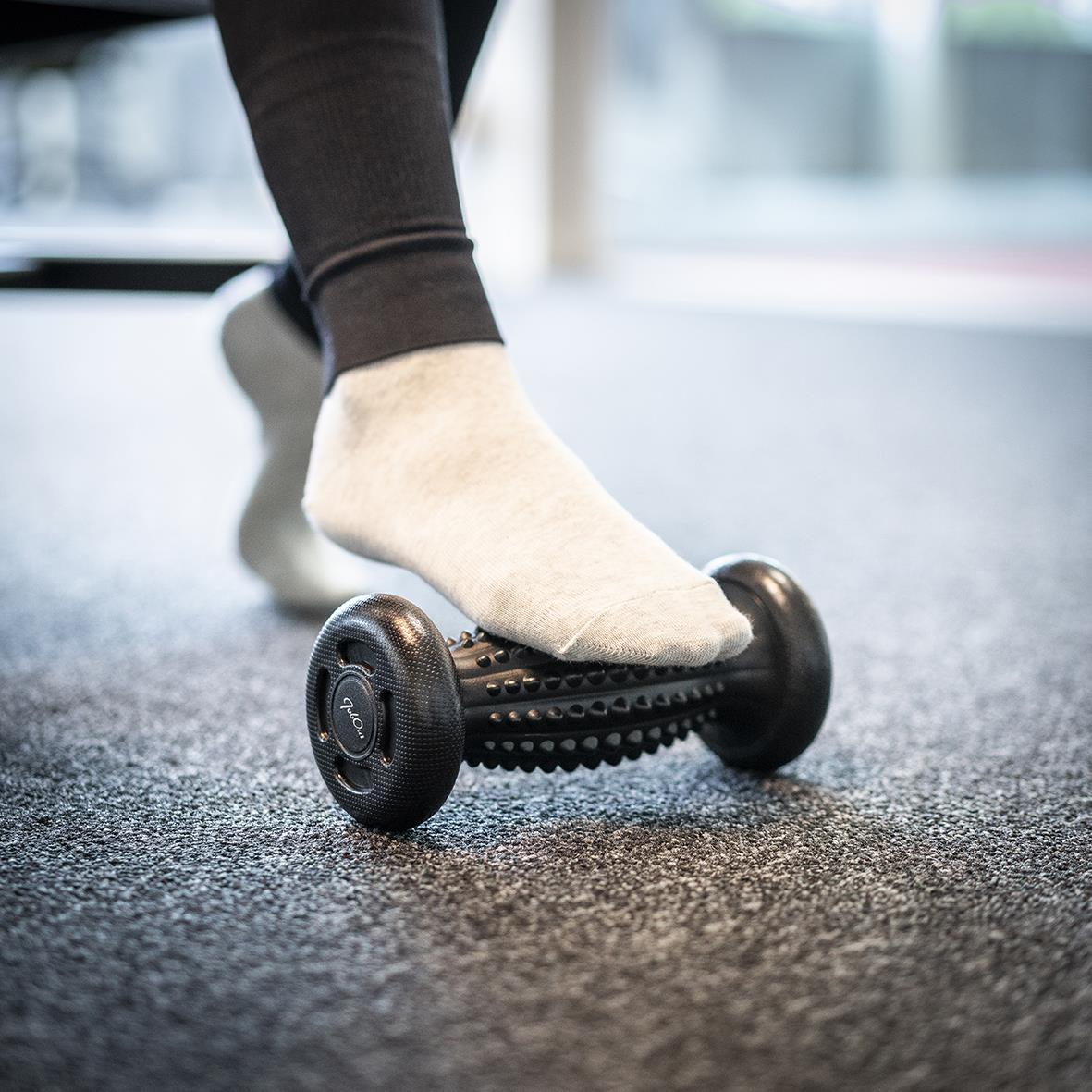 Person använder en fotmassagerulle på golvet, klädd i sockor och svarta byxor