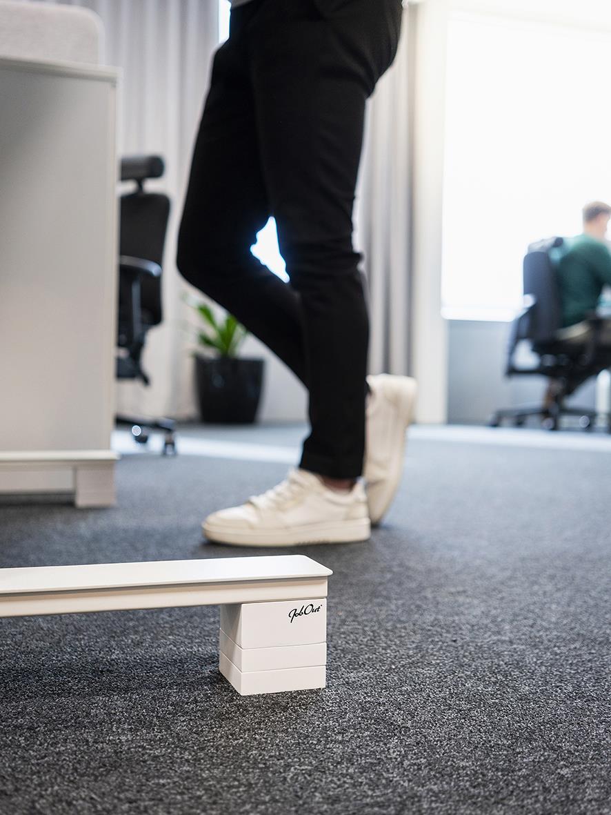 Person står vid skrivbordshöjare i kontorsmiljö, syns endast ben och fötter, svart byxa och vita sneakers