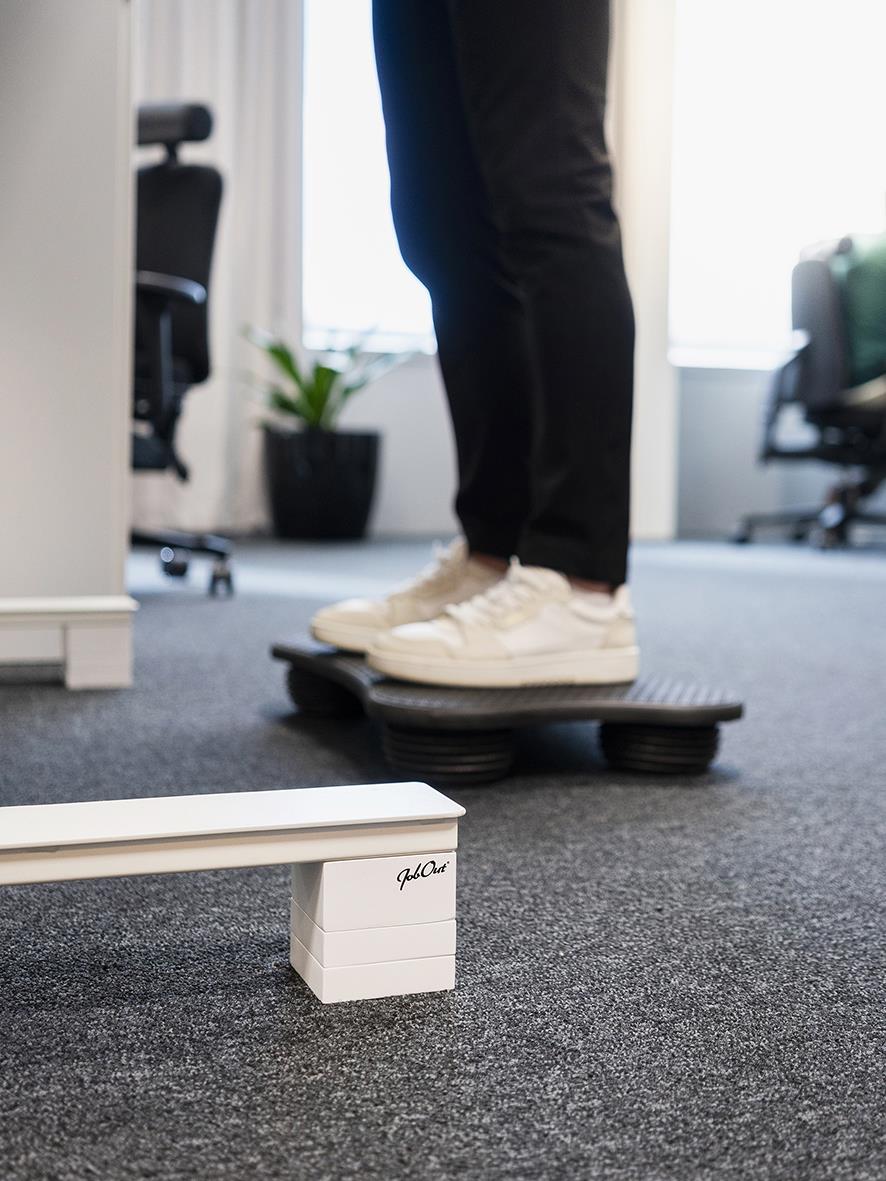 Person står på en svart skrivbordshöjare i ett kontor med grå matta och kontorsmöbler i bakgrunden