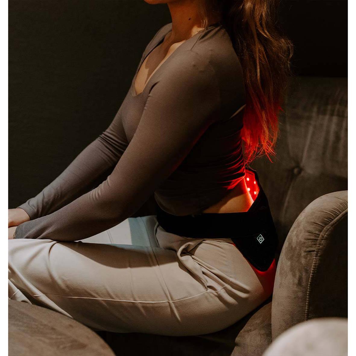 Kvinna använder ett infrarött ljusbälte för hållningstöd sittande på en soffa i ett mörkt rum