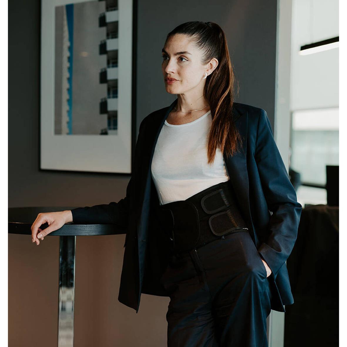 Kvinna bär hållningsväst i ett kontorsmiljö, klädd i vit t-shirt och mörk blazer