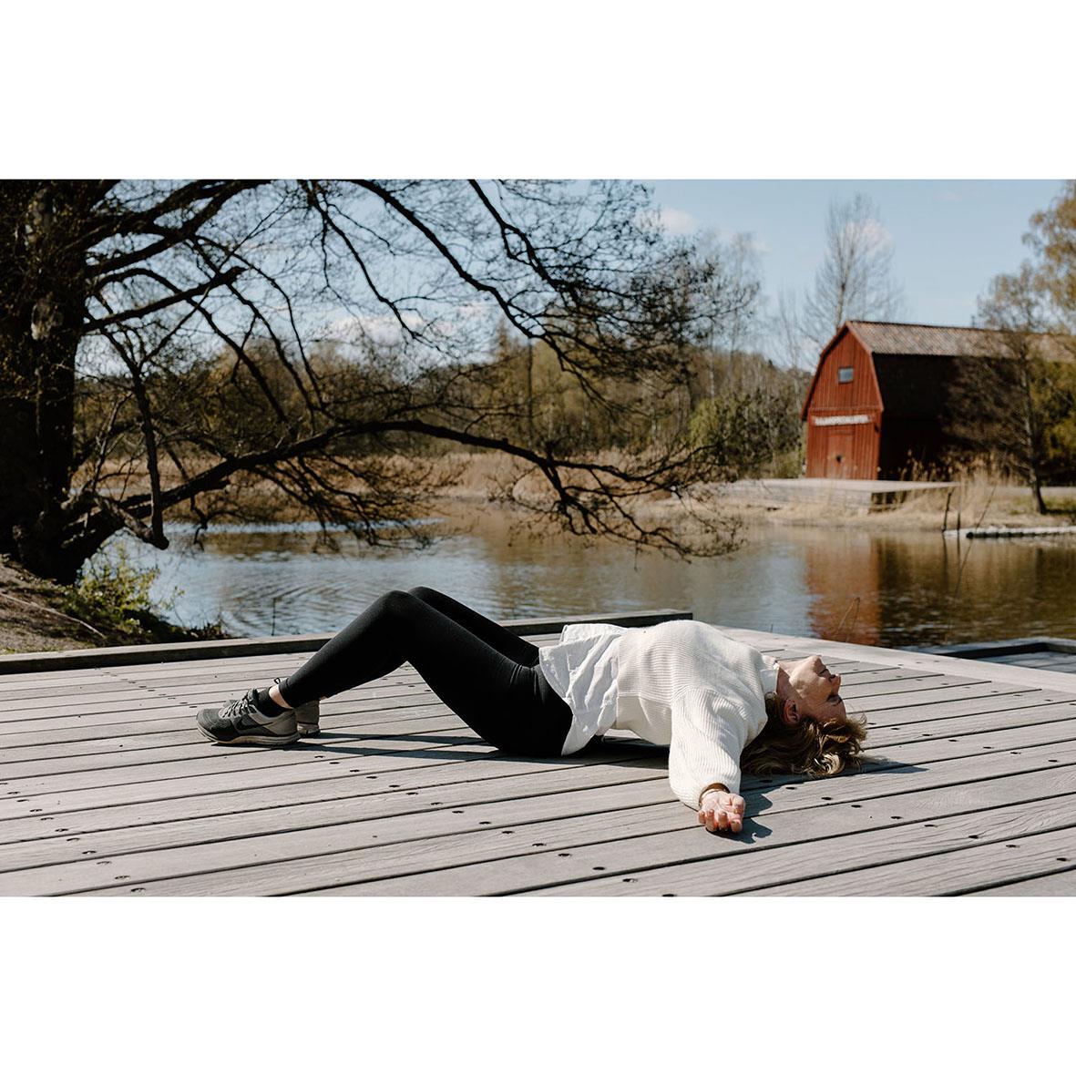 Kvinna utför ryggsträckning på trädäck utomhus med sjö och röd lada i bakgrunden