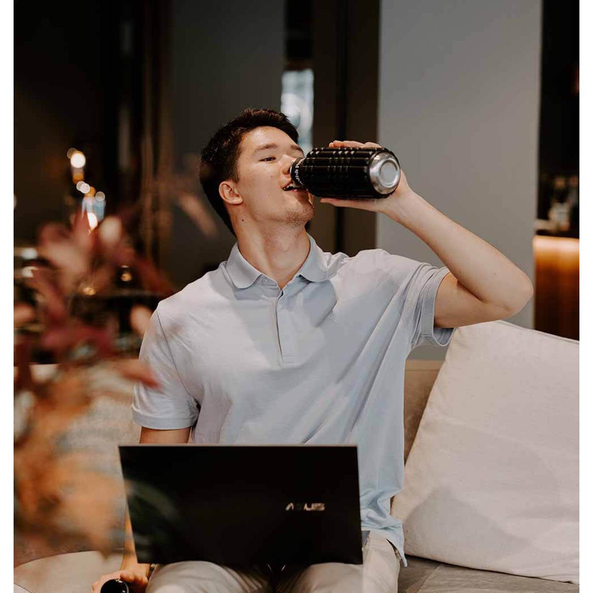 Ung man dricker vatten från vattenflaska medan han använder laptop i ett vardagsrum