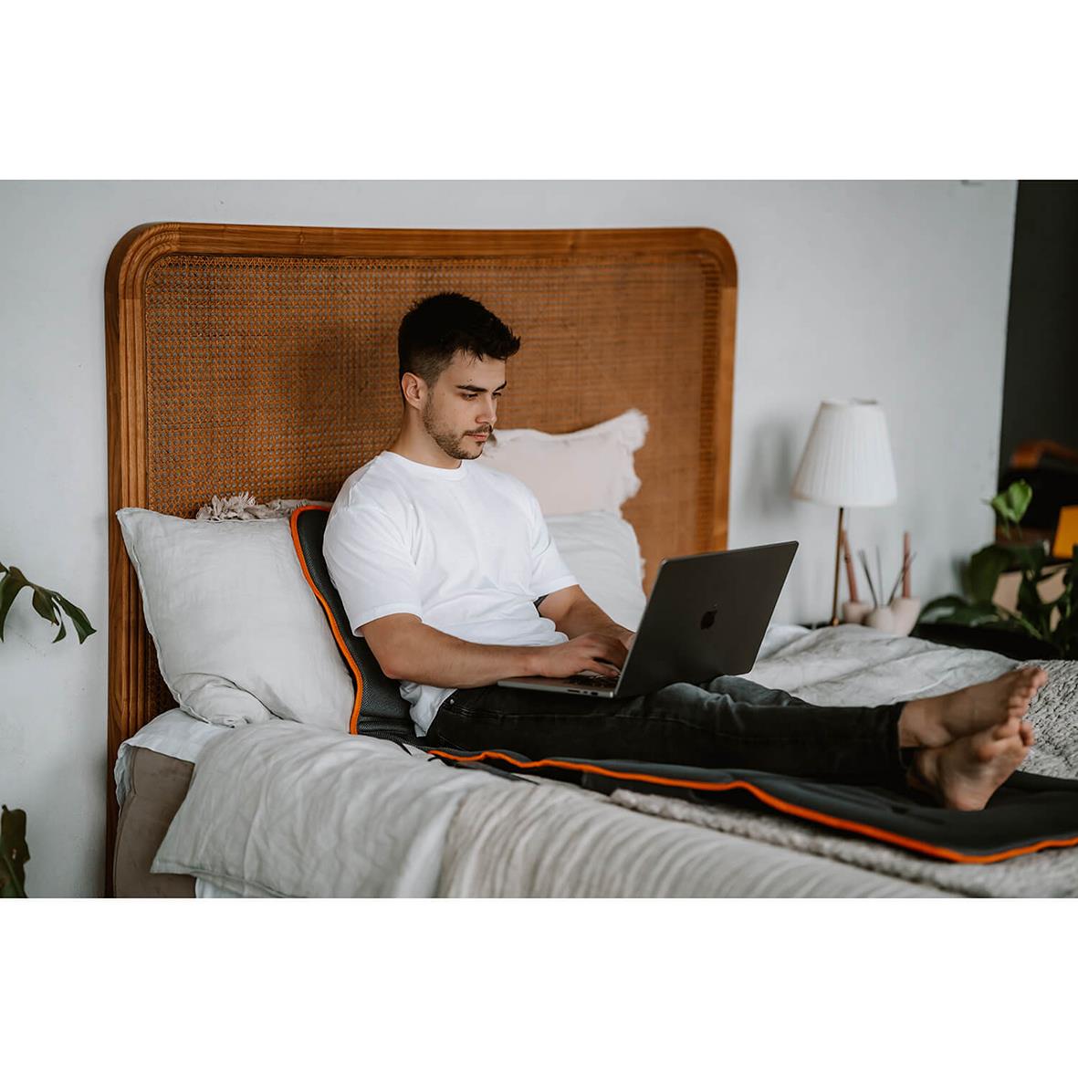 Man ligger på en massagematta i sängen, använder en laptop, i ett rum med växter och en vit lampa