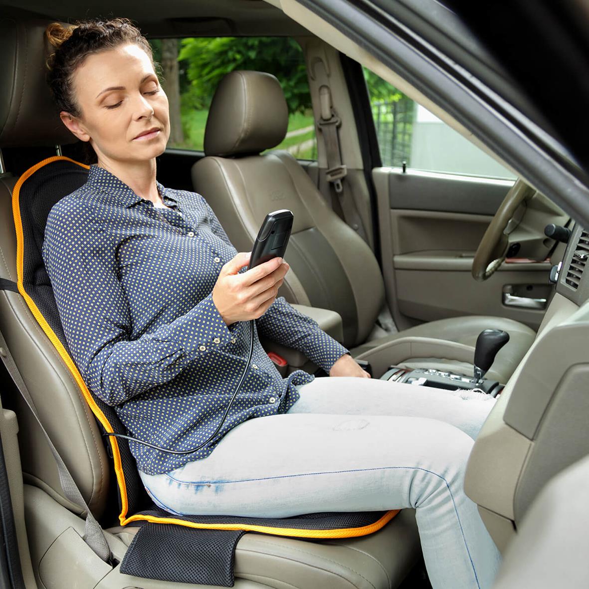 Kvinna sitter i bilens förarsäte med Medivon Cosy Mesh massagesits, kopplar av och använder mobiltelefonen