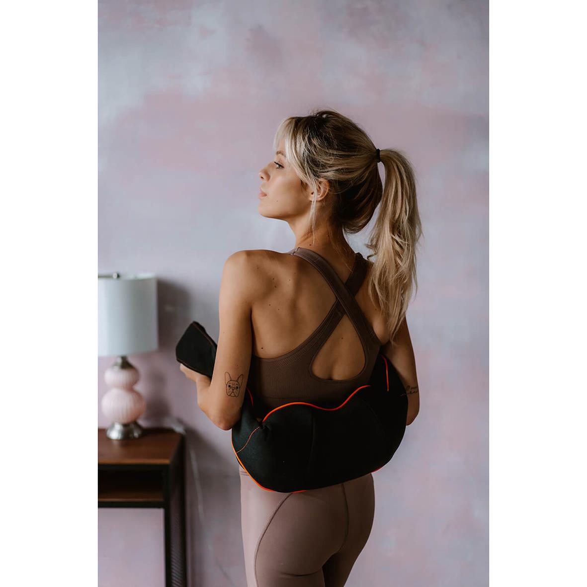 Kvinna med svart nackmassageapparat Medivon Simple bär den på ryggen, står vid vägg i rum med rosa vägg och nattduksbord