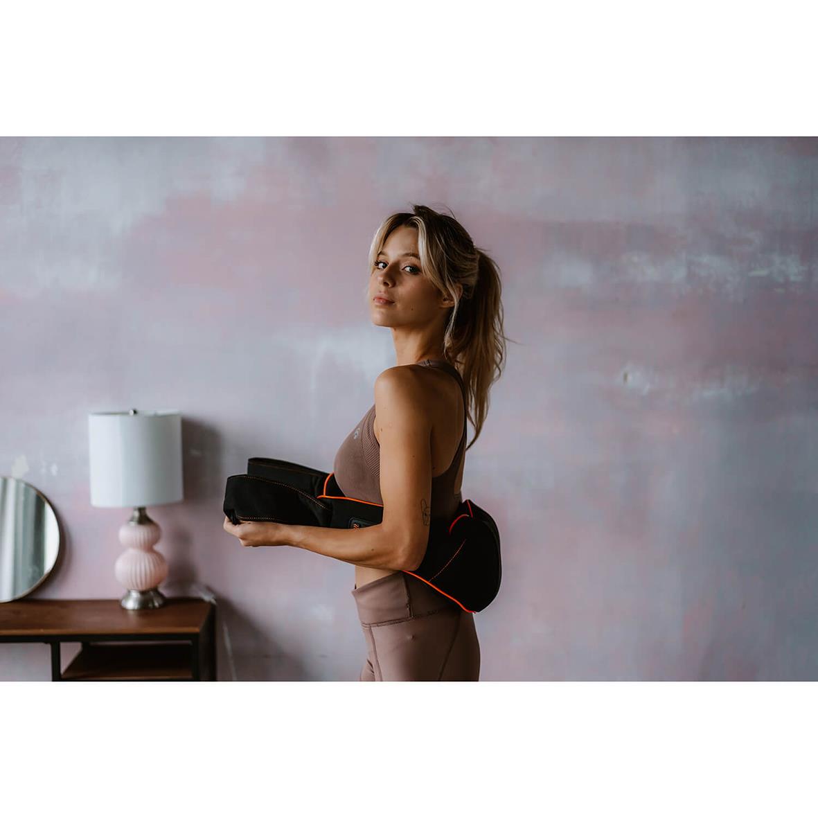 Kvinna med nackmassageapparat i svart, står i ett rum med vägg och nattduksbord