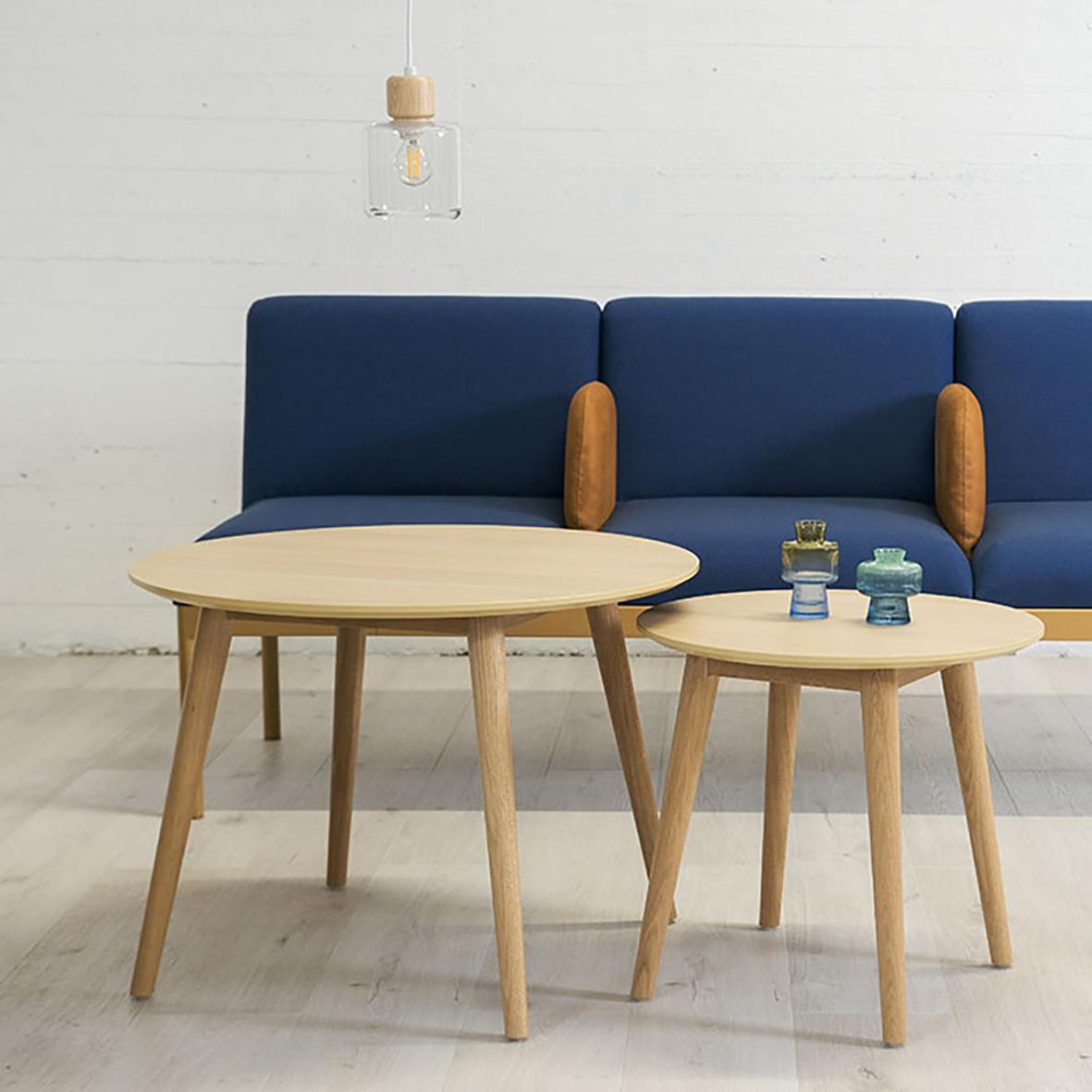 Två runda träbord framför en blå soffgrupp med träarmstöd i ett minimalistiskt vardagsrum