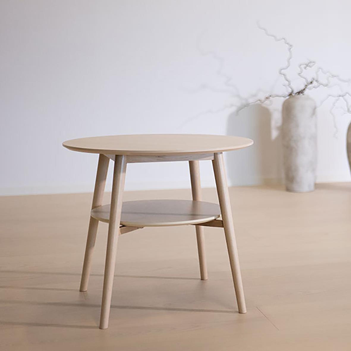 Litet runt träbord med två nivåer står på ett ljust trägolv i ett minimalistiskt rum med en stor vas med kvistar i bakgrunden