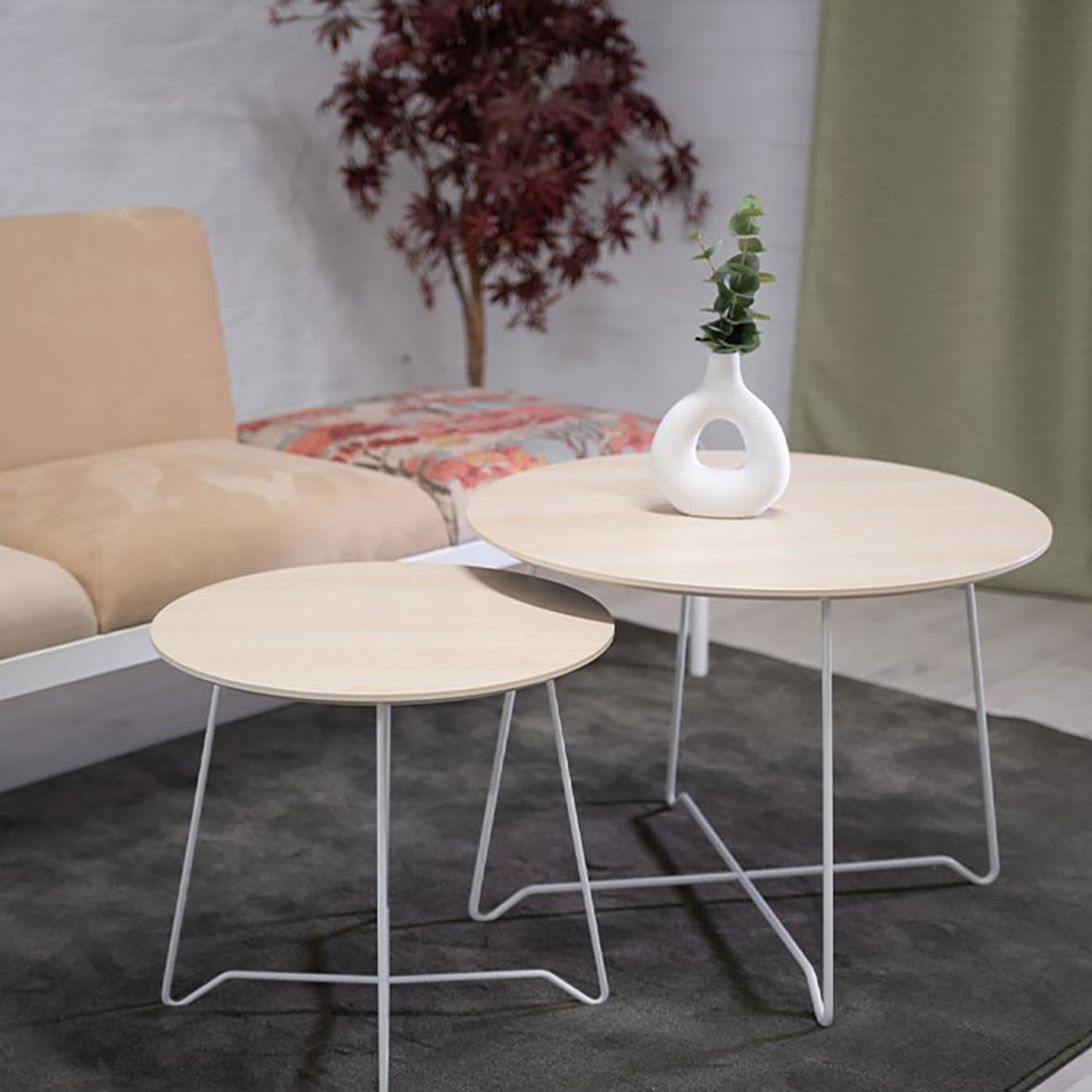 Två runda soffbord med vita metallben, ett större och ett mindre, på en mörk matta i ett vardagsrum med beige soffa och växt i bakgrunden