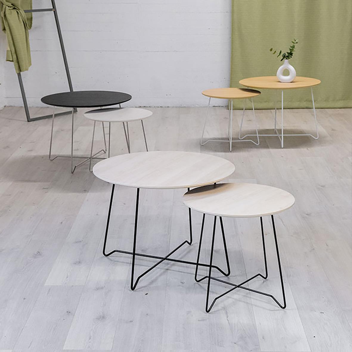 Två vita runda soffbord med svarta och vita metallben, ett mindre och ett större, i ett inomhusmiljö med ljus golv och gröna och gula gardiner