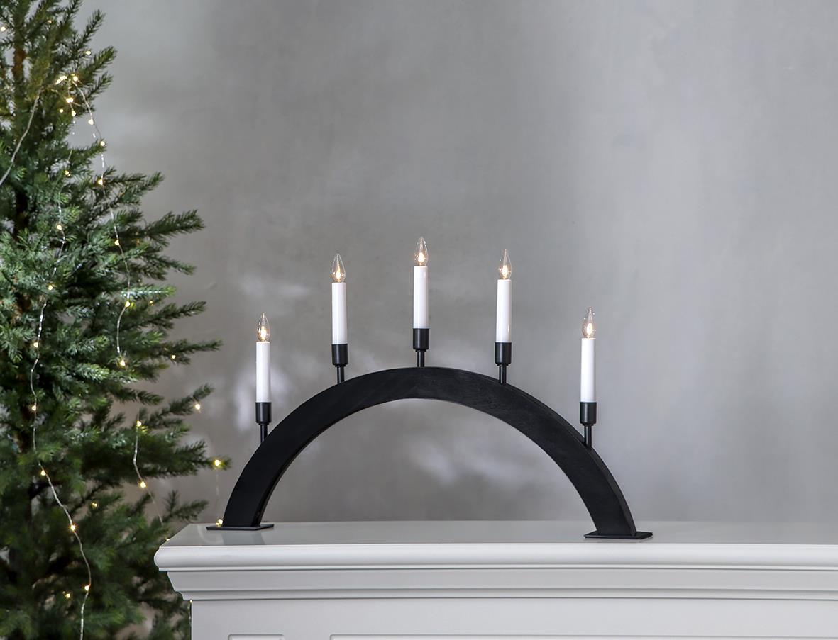 Svart ljusstake i bågform med fem vita ljus, 57x38cm, placerad på en vit möbel framför en grå vägg och julgran